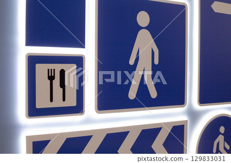 Various directional and informational signs. Symbols include pedestrian icon, dining symbol with utensils, and directional arrow, all set on blue background with illuminated edges 129833031