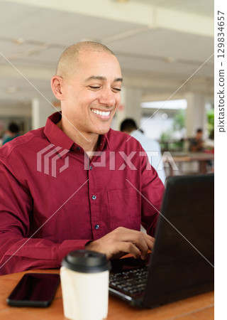 Bald man working on laptop computer at outdoor coffee shop in business or campus setting 129834657
