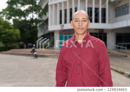 Confident bald Italian man in burgundy shirt standing in front of university building on sunny day Confident bald Italian man in burgundy shirt standing in front of university building on sunny day 129834663