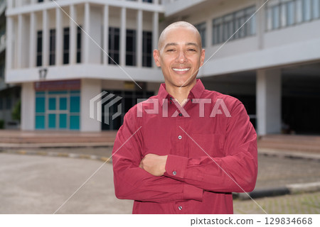 Confident bald Italian man in burgundy shirt standing in front of university building on sunny day 129834668
