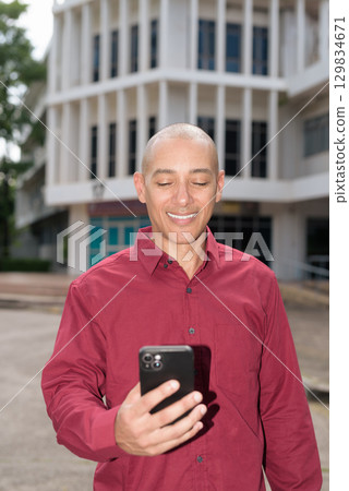 Bald man using smartphone while standing outside university building 129834671