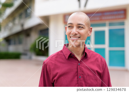 Confident bald Italian man in burgundy shirt standing in front of university building on sunny day Confident bald Italian man in burgundy shirt standing in front of university building on sunny day 129834678