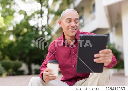 Bald man using digital tablet while sitting outdoors with coffee on stairs Bald man using digital tablet while sitting outdoors with coffee on stairs 129834685