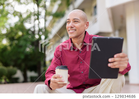 Bald man using digital tablet while sitting outdoors with coffee on stairs Bald man using digital tablet while sitting outdoors with coffee on stairs 129834686