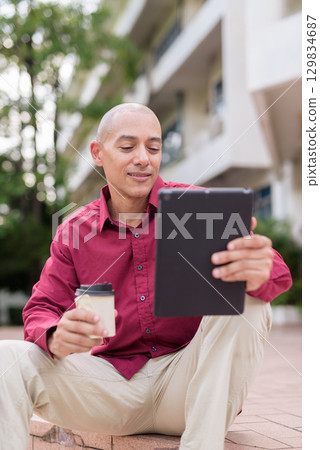 Bald man using digital tablet while sitting outdoors with coffee on stairs Bald man using digital tablet while sitting outdoors with coffee on stairs 129834687