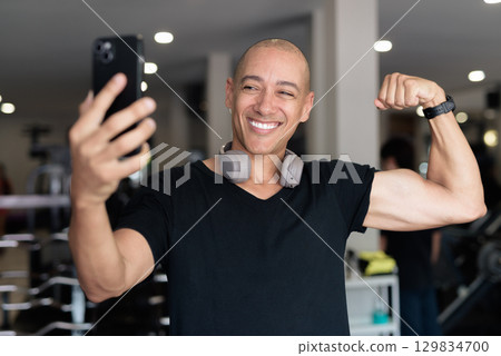 Hispanic bald man athlete in gym using mobile phone 129834700