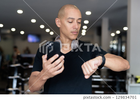 Hispanic bald man athlete in gym using smartwatch fitness tracker 129834701