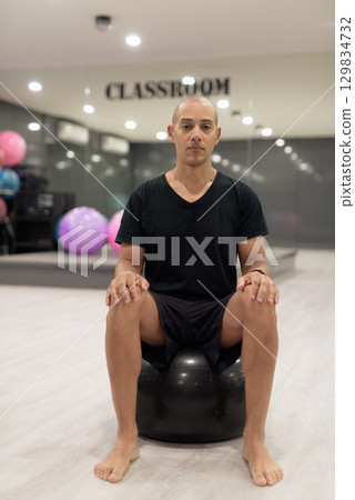 Hispanic bald man athlete in gym doing Pilates workout with exercise ball 129834732