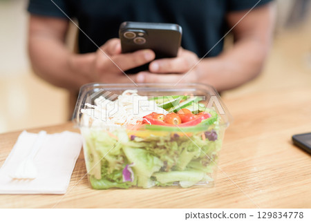 Hand holding smartphone over healthy salad in mall food court, indoor lifestyle detail 129834778