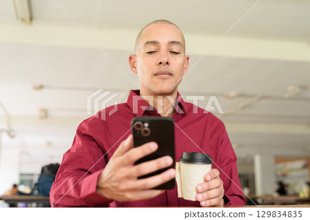 Man using mobile phone taking selfie at outdoor cafe in university area with takeaway coffee 129834835