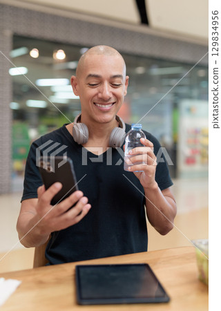 Man using tablet while eating salad and drinking water in mall cafe 129834956