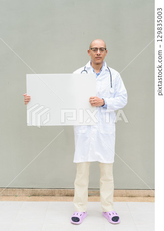 Full Length Portrait of Male Doctor in Lab Coat with Stethoscope Holding Placard with Copy Space 129835003