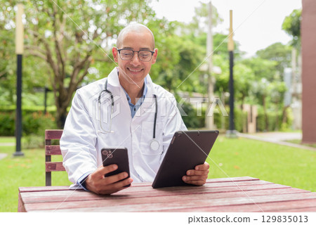 Doctor in uniform relaxing outdoors in hospital garden, smiling and confident using tablet computer Doctor in uniform relaxing outdoors in hospital garden, smiling and confident using tablet computer 129835013