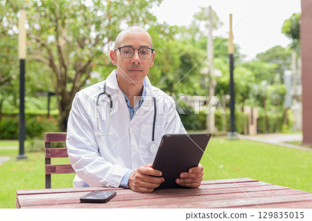 Doctor in uniform relaxing outdoors in hospital garden, smiling and confident using tablet computer Doctor in uniform relaxing outdoors in hospital garden, smiling and confident using tablet computer 129835015
