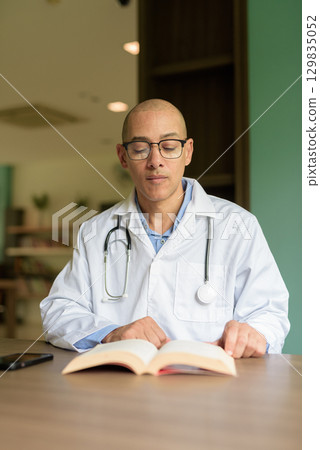 Doctor Sitting Indoors in Clinic or Hospital Area Library with Stethoscope Reading Book 129835052
