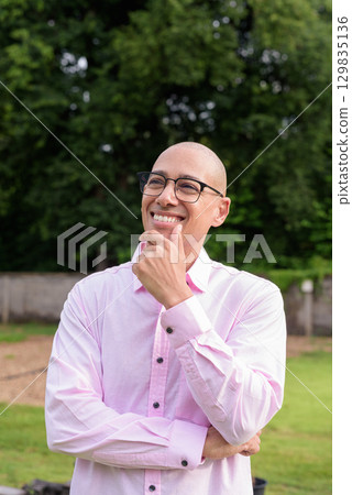 Bald Hispanic man thinking and looking up with hand on chin outdoors 129835136