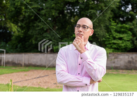 Bald Hispanic man thinking and looking up with hand on chin outdoors 129835137