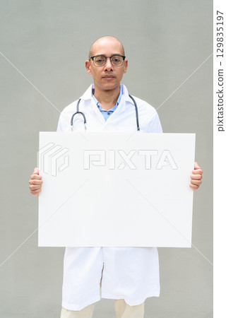 Hispanic Male Doctor in Lab Coat with Stethoscope on Plain Background Holding Empty Placard Hispanic Male Doctor in Lab Coat with Stethoscope on Plain Background Holding Empty Placard 129835197
