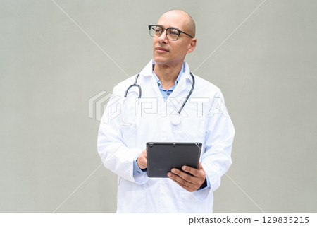 Hispanic Male Doctor in Lab Coat with Stethoscope on Plain Background Using Tablet Computer Hispanic Male Doctor in Lab Coat with Stethoscope on Plain Background Using Tablet Computer 129835215