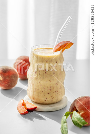 Smoothie with peach and banana in a glass on a white table near window with tropical fruit slices 129836435