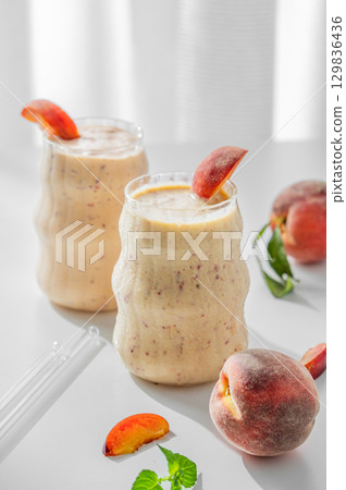 Two glasses of smoothie with peach and banana on a light background near window with tropical fruits 129836436