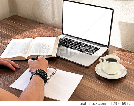A young man with short black hair studies at a wooden table. A laptop, an open book, and a cup of coffee are present.  129836932