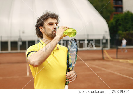 Portrait of tennis player man, front view. Man standing on a court and drinking a water from a sportive bottle. Man wearing yellow t-shirt. 129839805