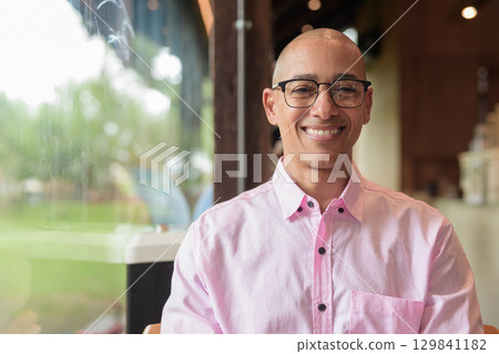Happy bald Hispanic man wearing pink casual business shirt and eyeglasses in coffee shop cafe 129841182