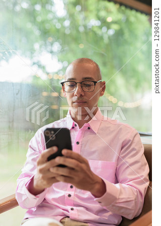 Bald Hispanic man wearing pink casual business shirt and eyeglasses in coffee shop using phone Bald Hispanic man wearing pink casual business shirt and eyeglasses in coffee shop using phone 129841201