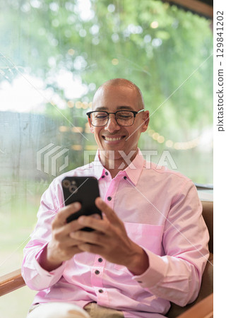 Bald Hispanic man wearing pink casual business shirt and eyeglasses in coffee shop using phone 129841202