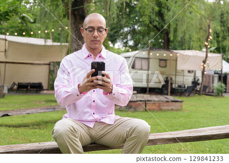 Bald Hispanic man wearing pink casual business shirt and eyeglasses outdoors using phone 129841233