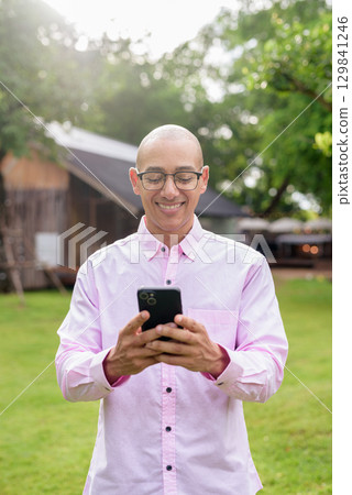 Bald Hispanic man wearing pink casual business shirt and eyeglasses outdoors using phone 129841246