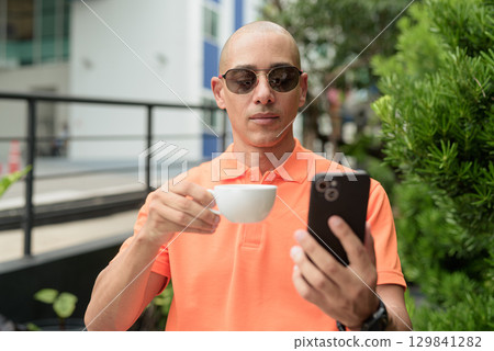 Bald Hispanic man face sitting outdoors at cafe restaurant wearing orange polo shirt using phone Bald Hispanic man face sitting outdoors at cafe restaurant wearing orange polo shirt using phone 129841282