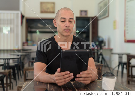 Bald Hispanic man sitting in outdoors cafe restaurant using digital tablet computer Bald Hispanic man sitting in outdoors cafe restaurant using digital tablet computer 129841313