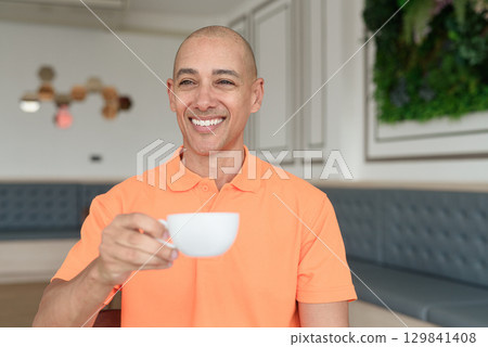 Happy man holding coffee cup inside cafe restaurant wearing orange polo shirt while looking away Happy man holding coffee cup inside cafe restaurant wearing orange polo shirt while looking away 129841408