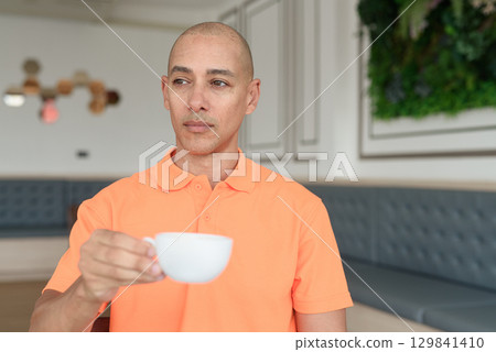 Man holding coffee cup inside cafe restaurant wearing orange polo shirt while thinking 129841410