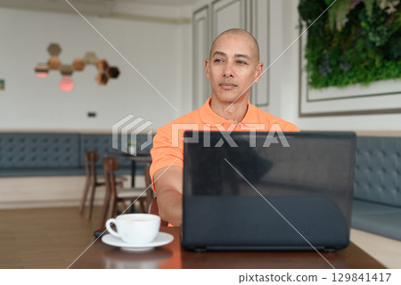 Bald Hispanic man smiling and using laptop computer in coffee shop restaurant while thinking 129841417
