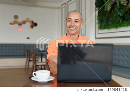 Bald Hispanic man smiling and using laptop computer in coffee shop restaurant while thinking 129841418