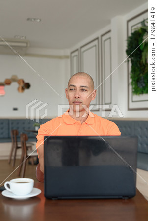 Bald Hispanic man smiling and using laptop computer in coffee shop restaurant while thinking 129841419