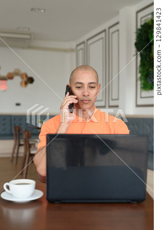 Bald Hispanic man using laptop computer in coffee shop restaurant while talking on phone 129841423