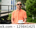 Bald Hispanic man face sitting outdoors at cafe restaurant wearing orange polo shirt 129841425