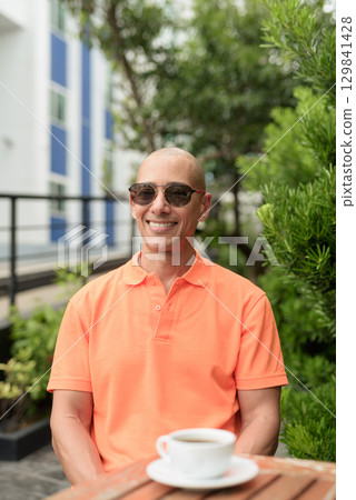 Bald Hispanic man face sitting outdoors at cafe restaurant wearing orange polo shirt 129841428