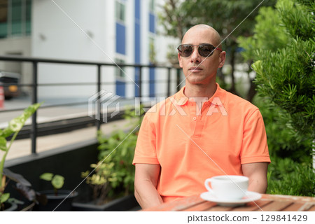 Bald Hispanic man face sitting outdoors at cafe restaurant wearing orange polo shirt 129841429