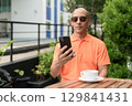 Bald Hispanic man face sitting outdoors at cafe restaurant wearing orange polo shirt using phone 129841431