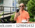 Bald Hispanic man face sitting outdoors at cafe restaurant wearing orange polo shirt using phone 129841432