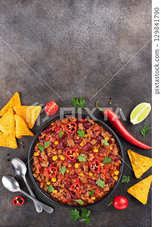 Chili con carne with fresh herbs and nachos chips on a gray background. Top view. Copy space. Mexican cuisine.  129841790