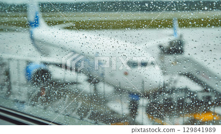 Raindrops blurring airport window, airplane resting on wet tarmac amid rainy weather conditions Raindrops blurring airport window, airplane resting on wet tarmac amid rainy weather conditions 129841989