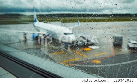 Raindrops blurring airport window view of parked airplane against gloomy runway backdrop 129842033