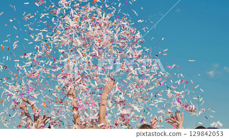 High school students joyfully celebrating graduation by tossing colorful confetti under a clear blue sky 129842053