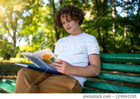 Curly-haired teenager sitting on the bench in the park and reading a book Curly-haired teenager sitting on the bench in the park and reading a book 129842295
